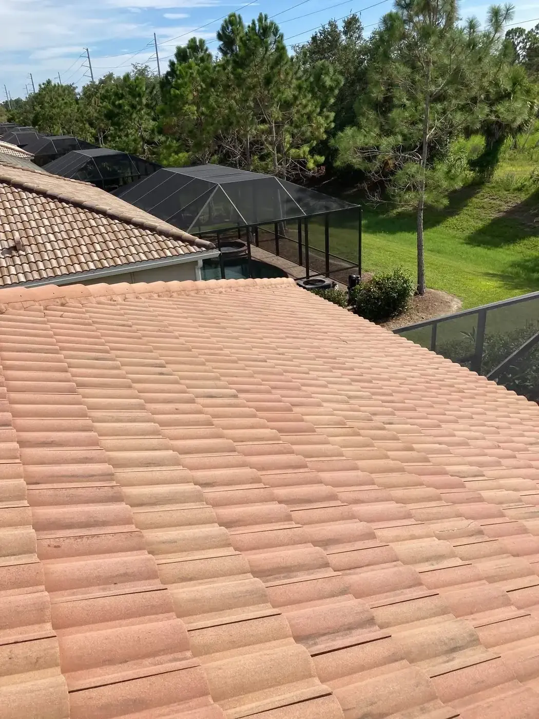 A barrel tile roof sloping down to a screened pool enclosure, more pool cages and pines behind it